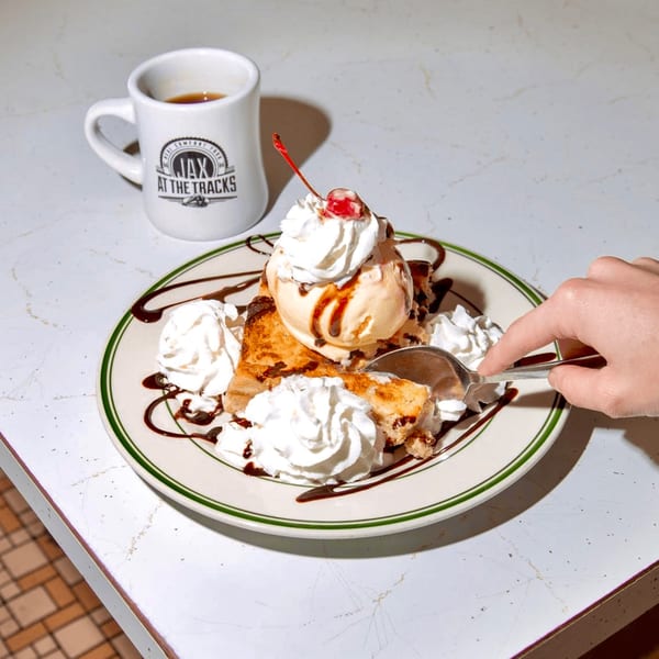 A hand spoons into a Jax At The Tracks dessert with ice cream, whipped cream, and cherry.
