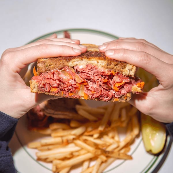 Hands holding a Jax At The Tracks pastrami sandwich with fries and a pickle spear.