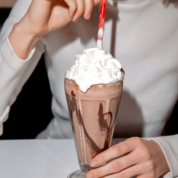 A person in a white sweater sips a chocolate milkshake with whipped cream and a red straw.