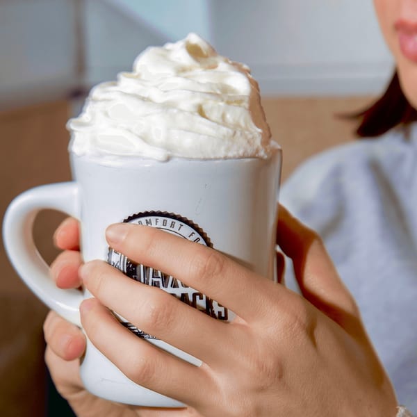 A person holds a white mug from Jax At The Tracks, topped with whipped cream.